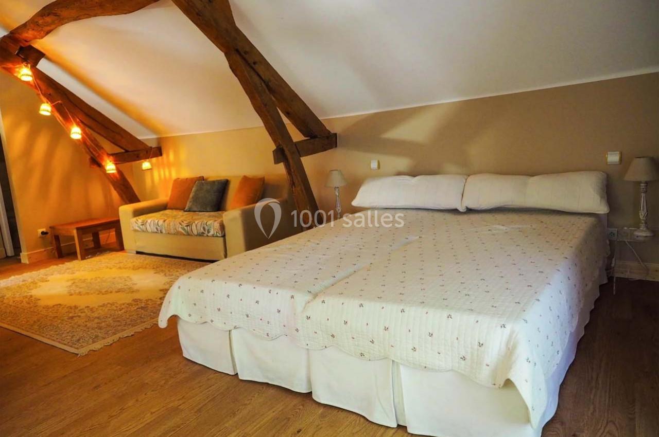 Chambre mansardée avec lit double, canapé, poutres apparentes et éclairage tamisé.