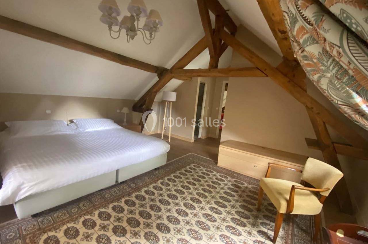 Chambre mansardée avec poutres apparentes, lit double, tapis décoratif, fauteuil et éclairage naturel.