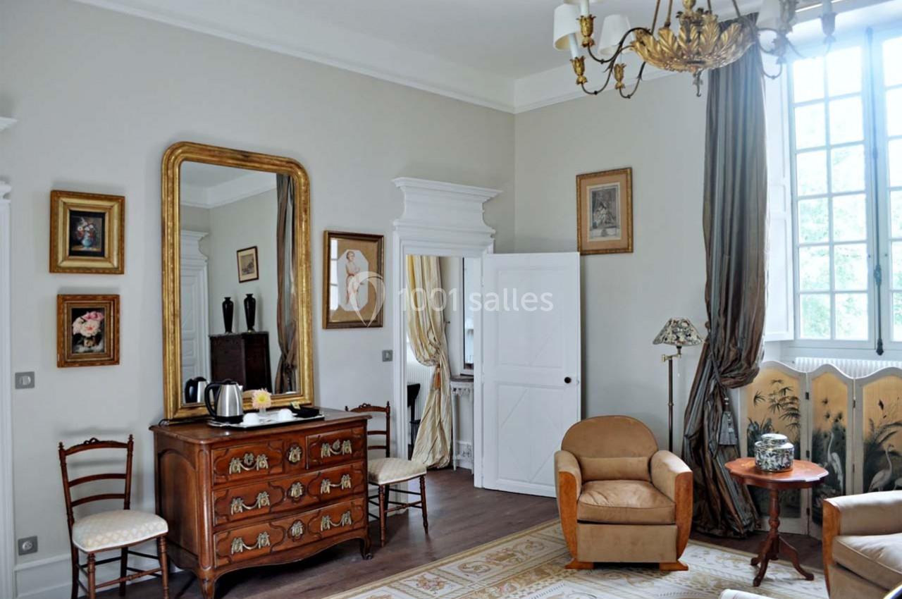 Chambre lumineuse avec mobilier classique, miroir doré, fauteuils, commode en bois et grande fenêtre à rideaux.