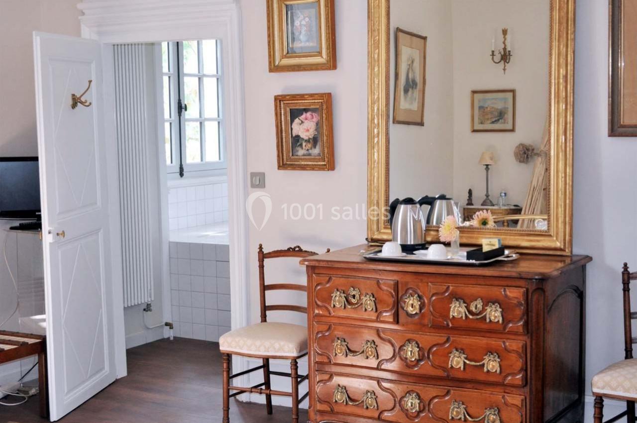 Commode en bois sculpté avec miroir doré dans une chambre lumineuse, vue sur une salle de bain carrelée blanche.