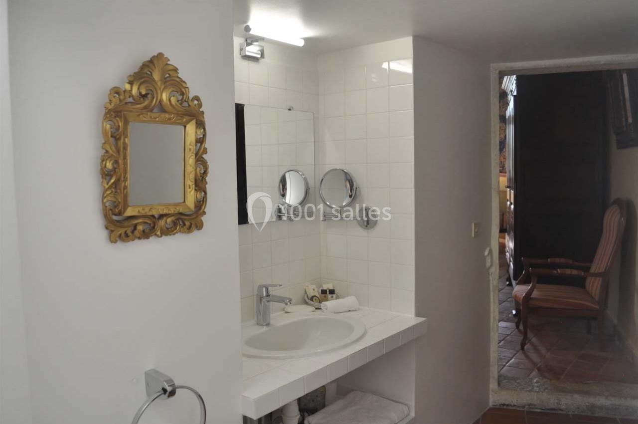 Salle de bain avec un lavabo, un miroir doré, des carreaux blancs et une ouverture donnant sur une pièce avec fauteuil.