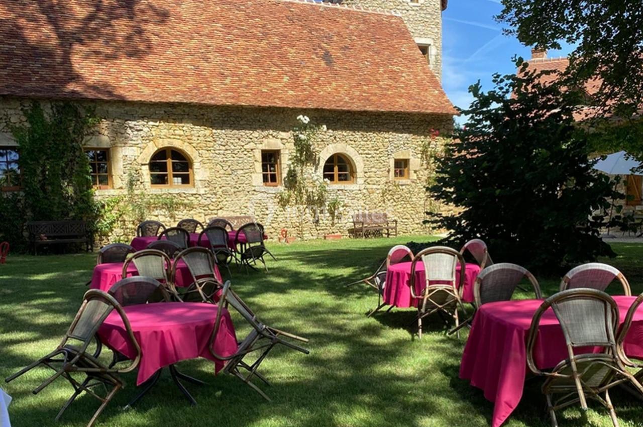 Tables rondes avec nappes roses disposées sur une pelouse, devant un bâtiment en pierre avec toit en tuiles.
