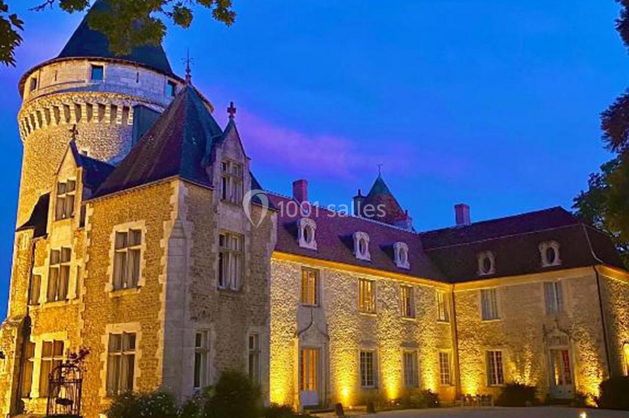 Façade d'un château en pierre éclairé de nuit, avec une tour ronde et un ciel bleu teinté de rose.