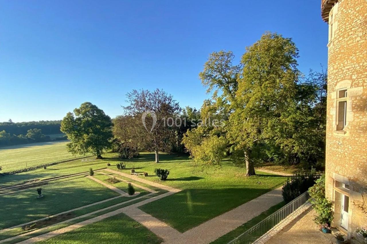 Vue d'un jardin avec allées gravillonnées, pelouse et arbres, bordé par un bâtiment en pierre sous un ciel bleu.