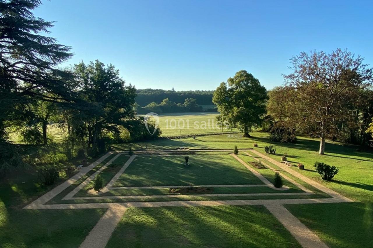 Vue sur un jardin géométrique avec allées en gravier, pelouse et arbres, bordé par une prairie sous un ciel dégagé.