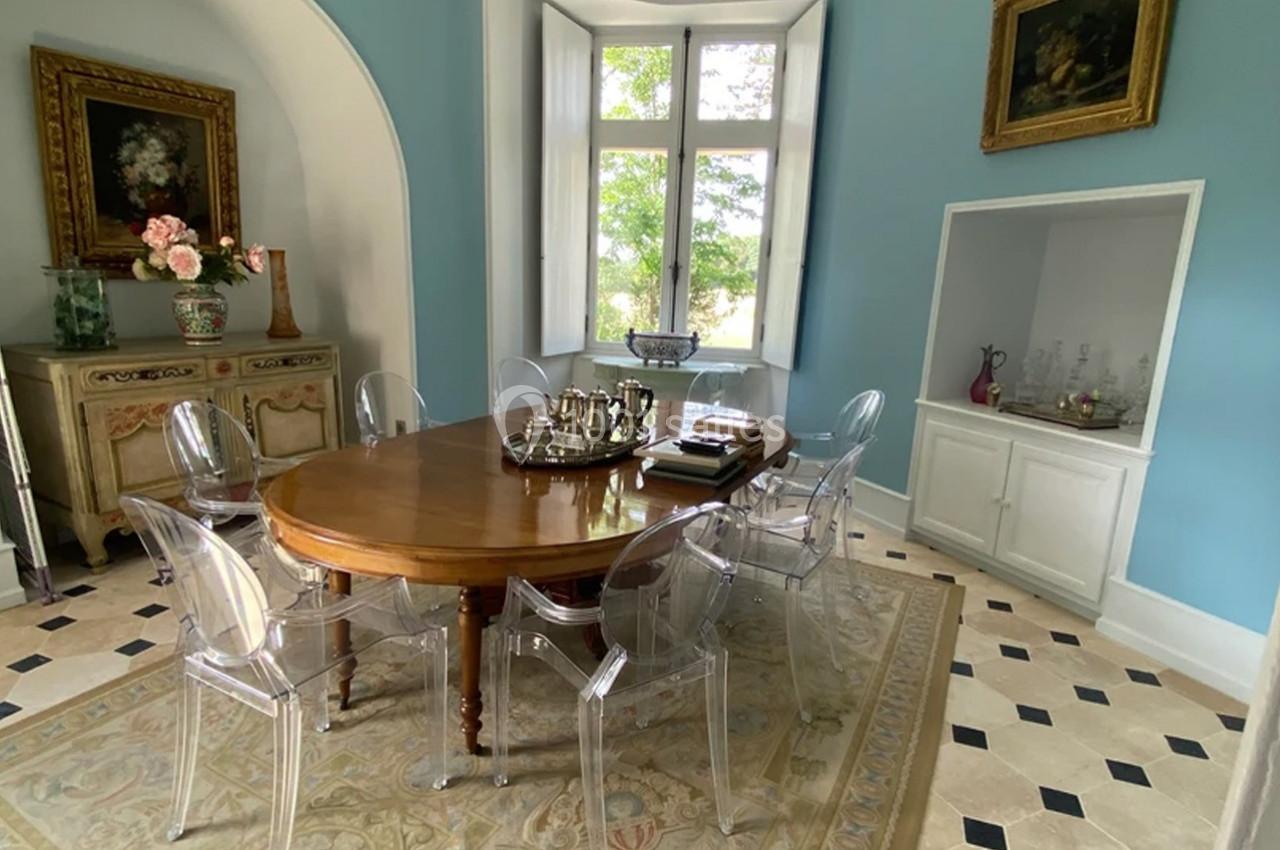 Salle à manger lumineuse avec table en bois ovale, chaises transparentes, murs bleus et vue sur un jardin par la fenêtre.