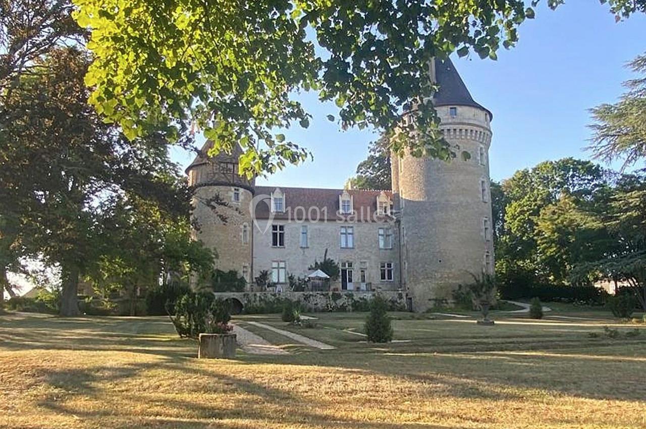 Château en pierre entouré d'arbres et d'une pelouse, avec une tour ronde et un ciel dégagé en arrière-plan.