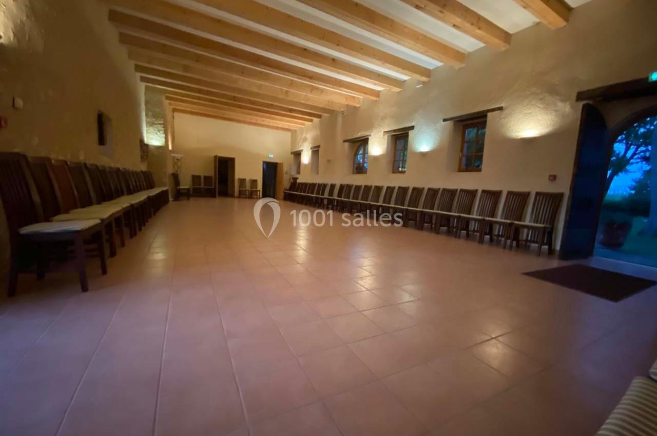 Grande salle vide avec un sol en carrelage, des murs clairs, un plafond en poutres apparentes et des chaises alignées le…