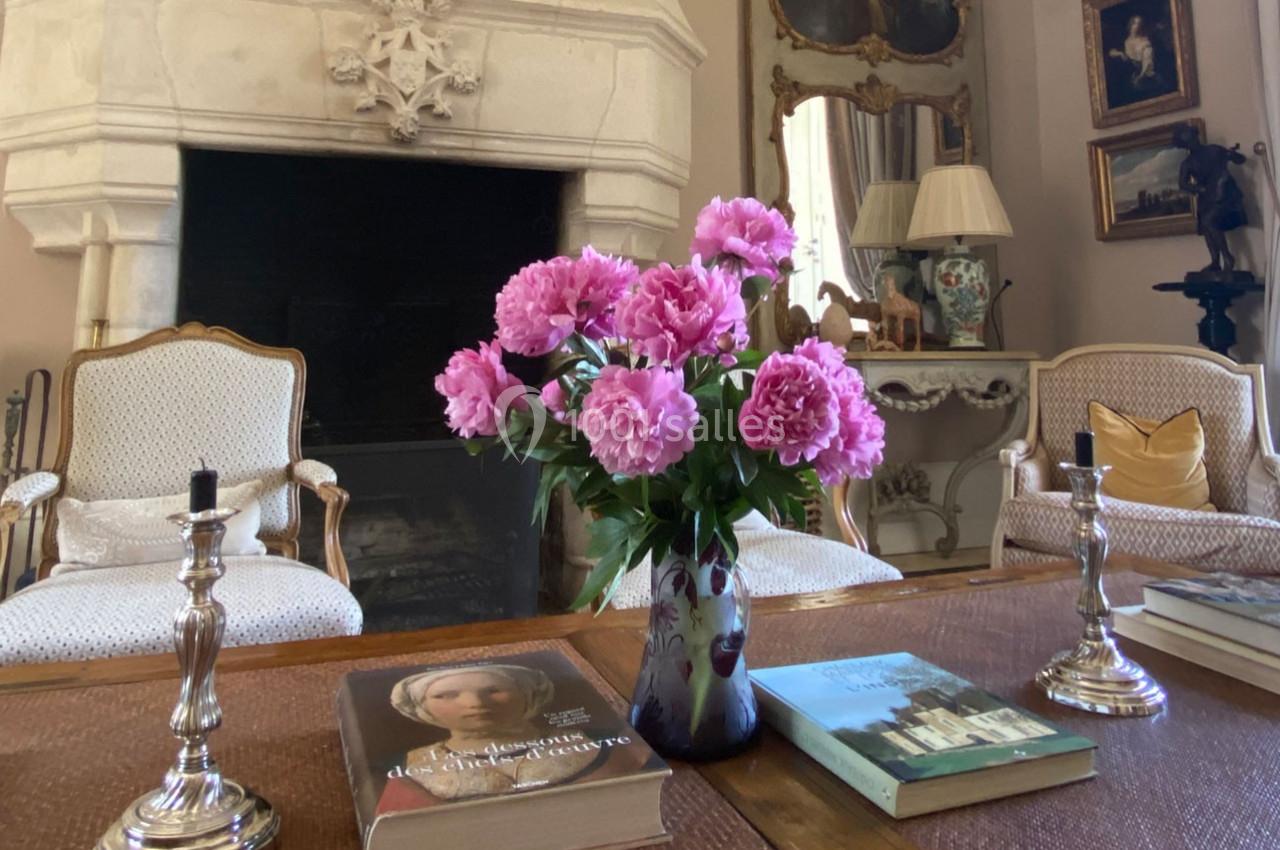 Vase de pivoines roses sur une table en bois, entouré de livres, chandeliers et fauteuils dans un salon élégant.