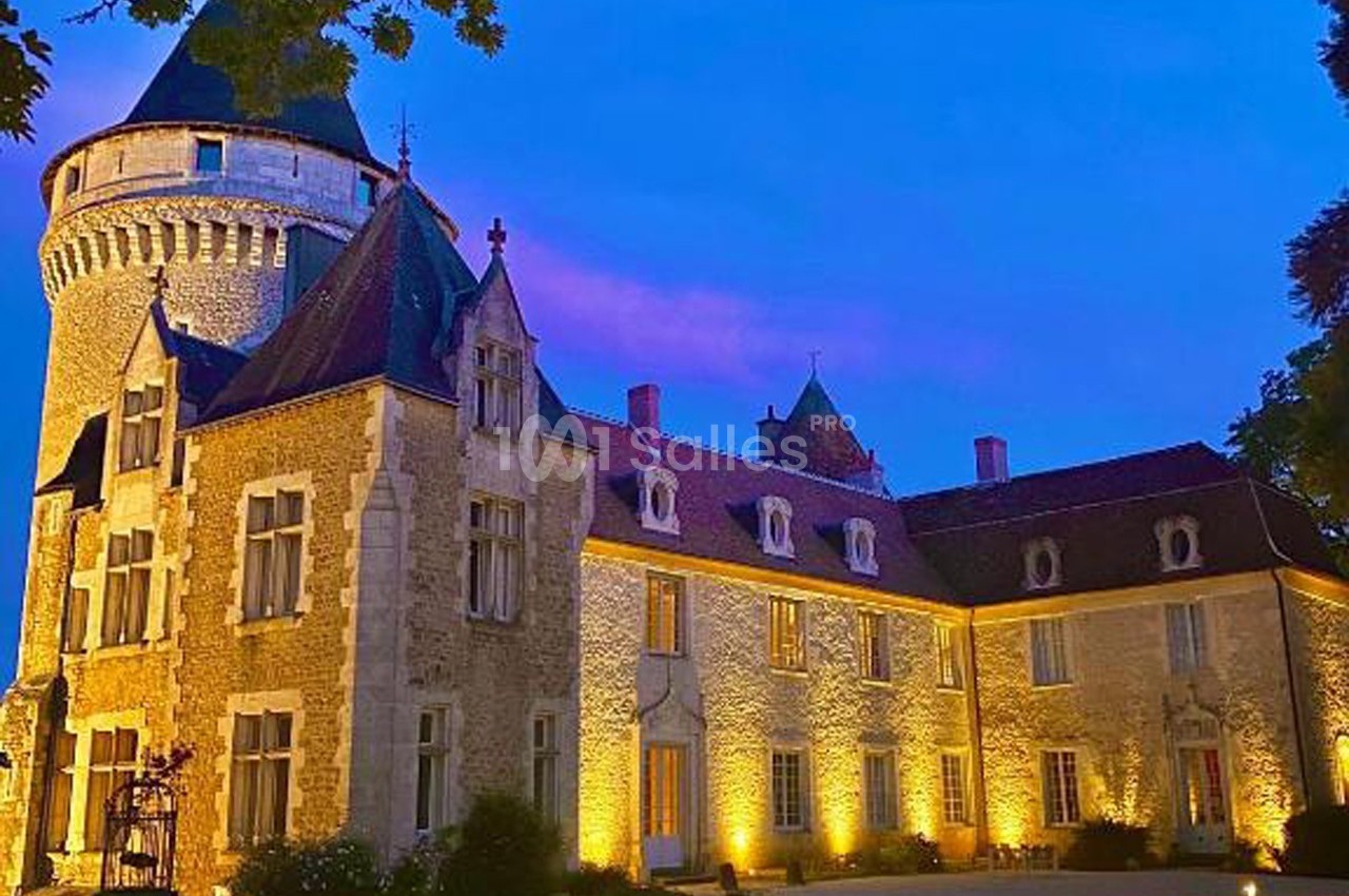 Façade d'un château en pierre éclairé de nuit, avec une tour ronde et un ciel bleu teinté de rose.