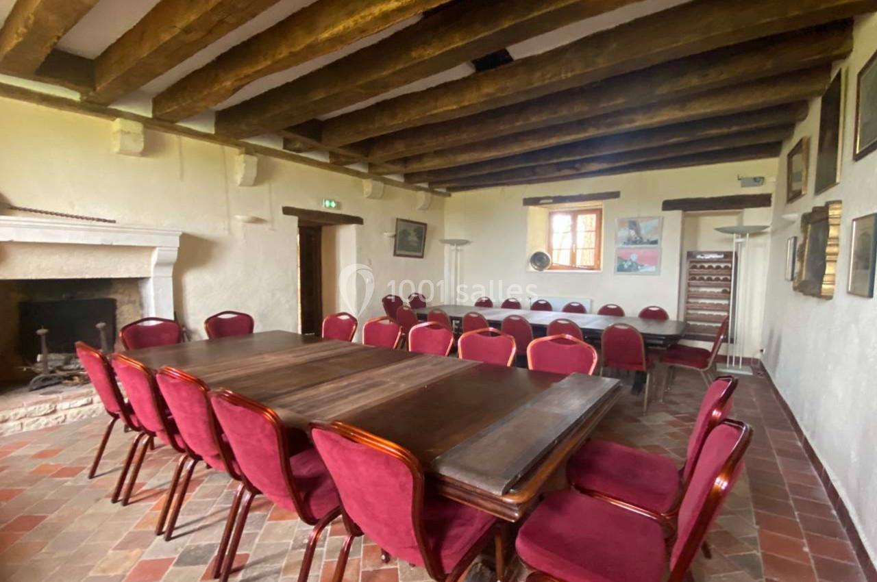 Salle avec poutres apparentes, tables en bois sombre et chaises rouges, sol en carrelage et cheminée en pierre.