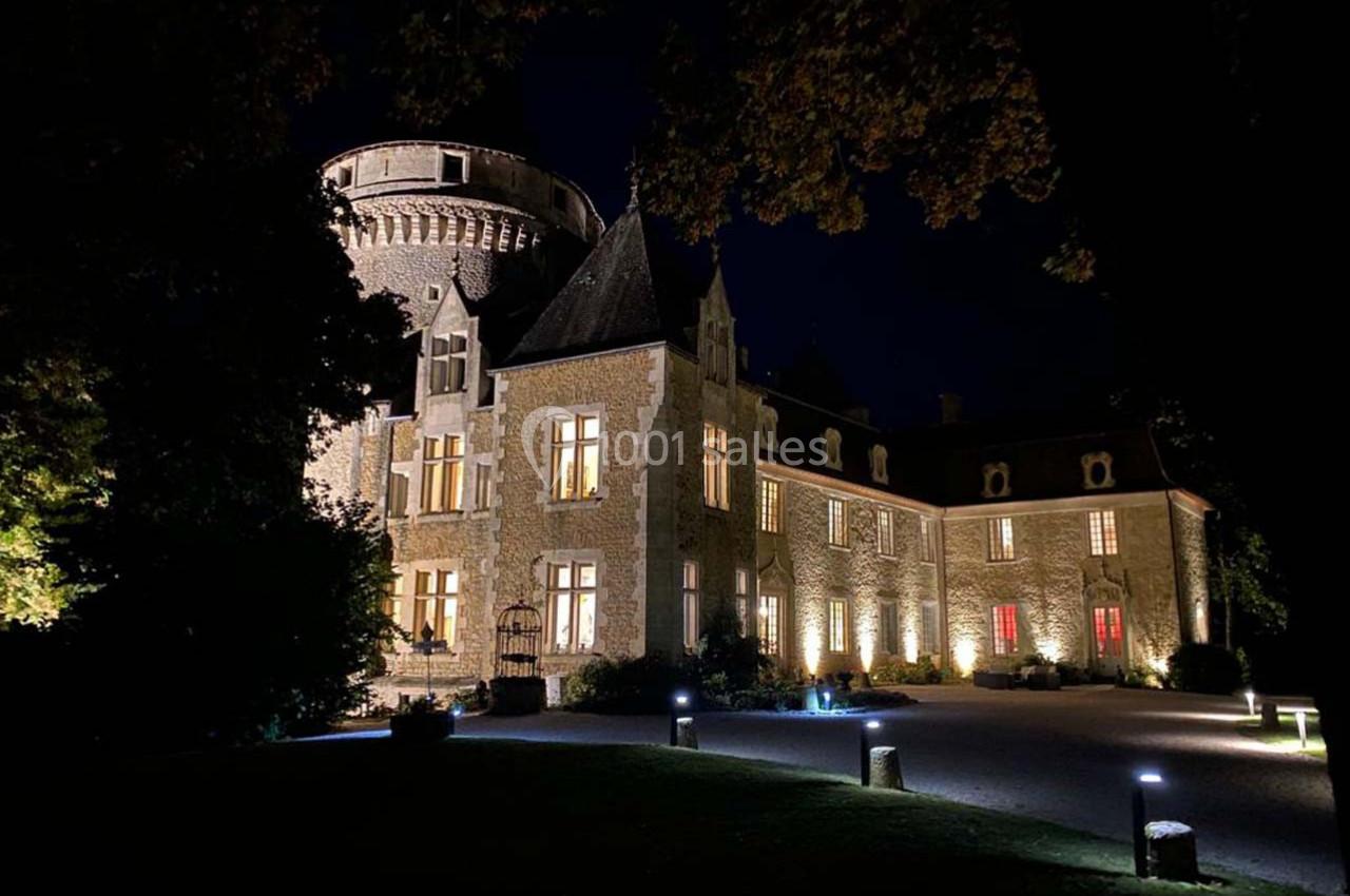 Château en pierre éclairé la nuit, entouré d'arbres et d'une allée illuminée.