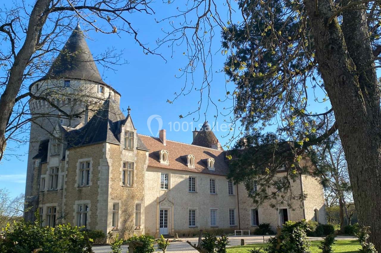 Château en pierre avec une tour ronde et un toit en ardoise, entouré d'arbres et d'un ciel dégagé.