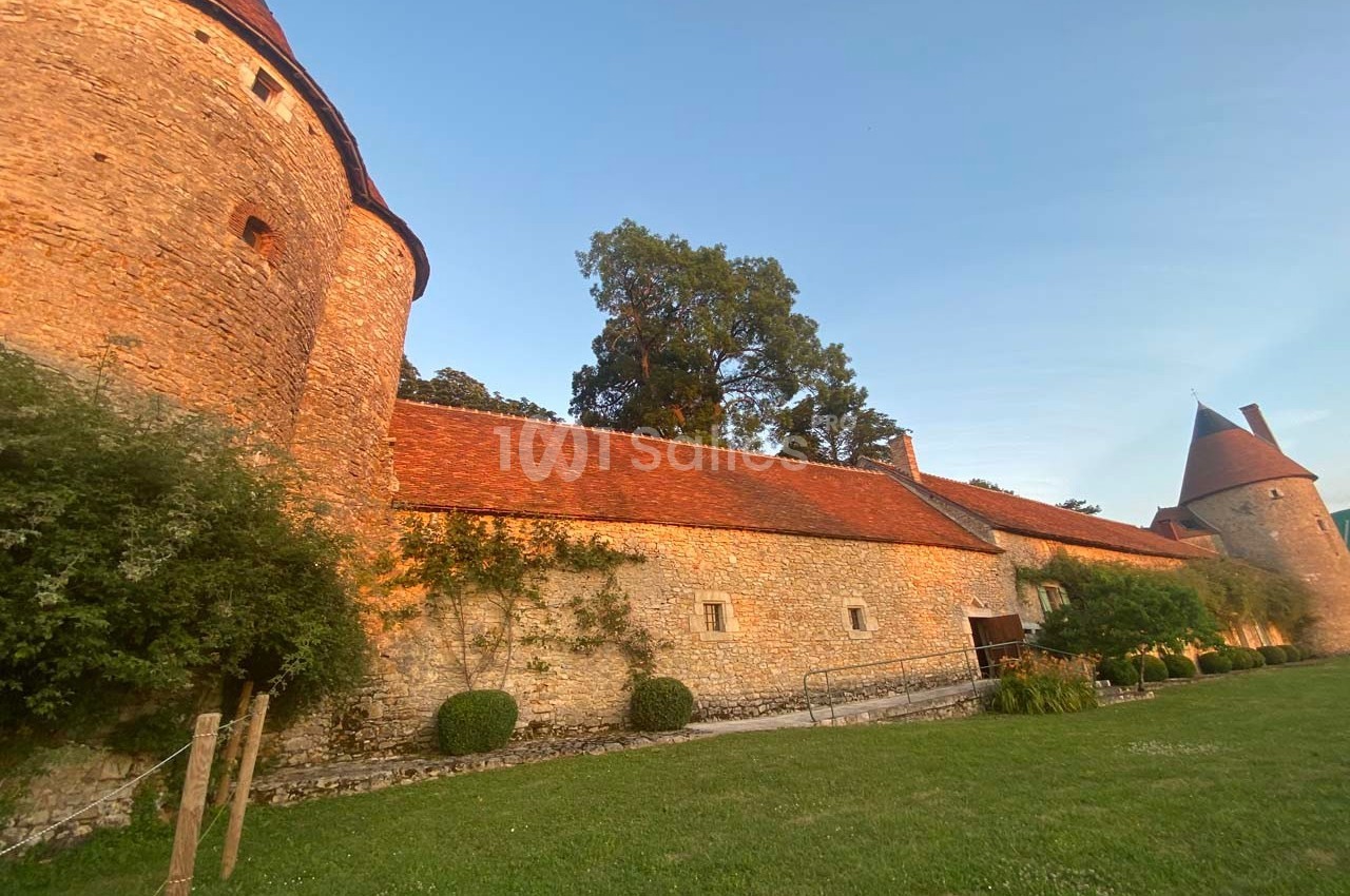 Mur en pierre d'un château avec tours rondes et toits en tuiles rouges, entouré de verdure au coucher du soleil.