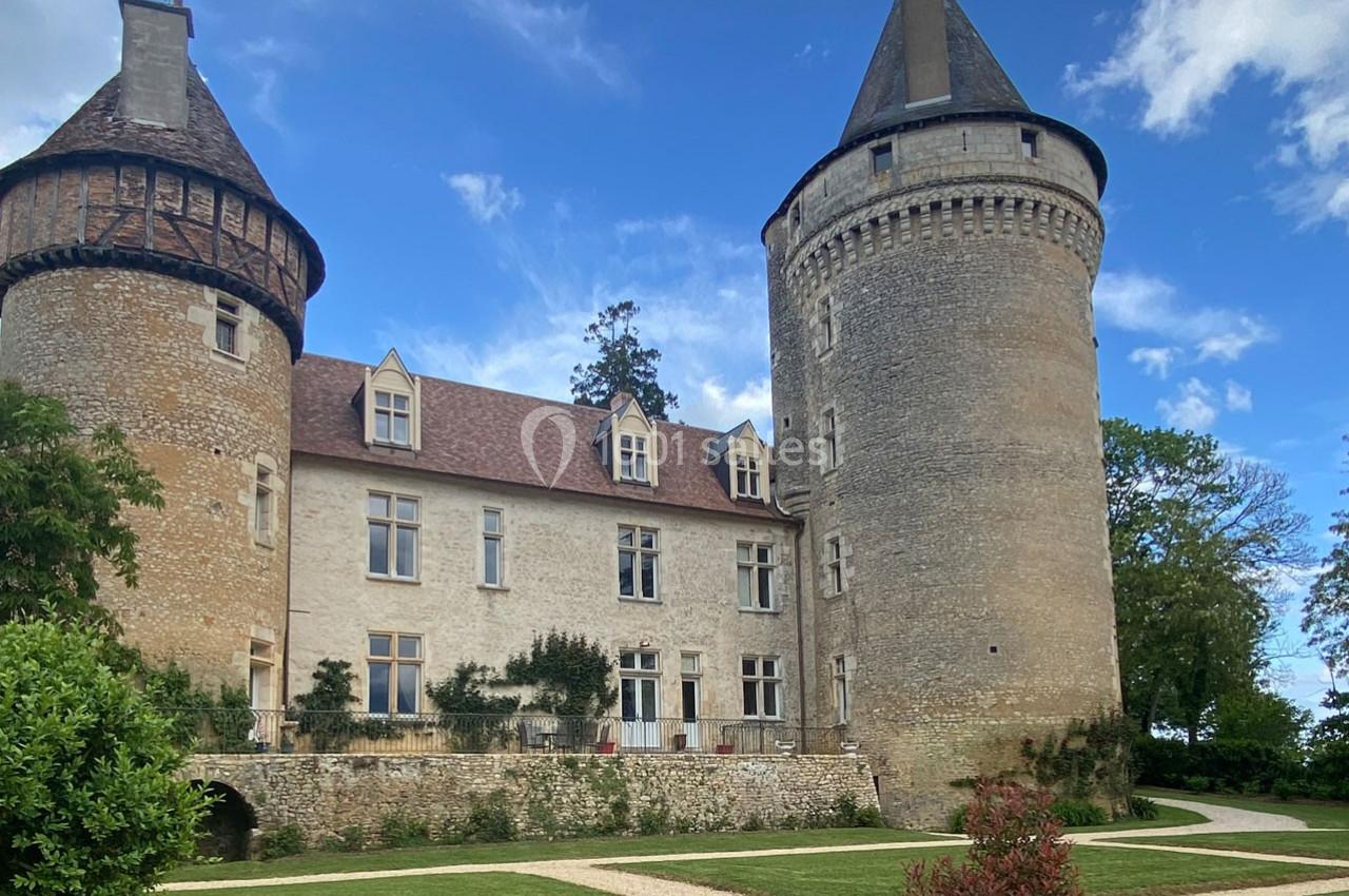 Château en pierre avec deux tours rondes, entouré de jardins et d'allées sous un ciel partiellement nuageux.