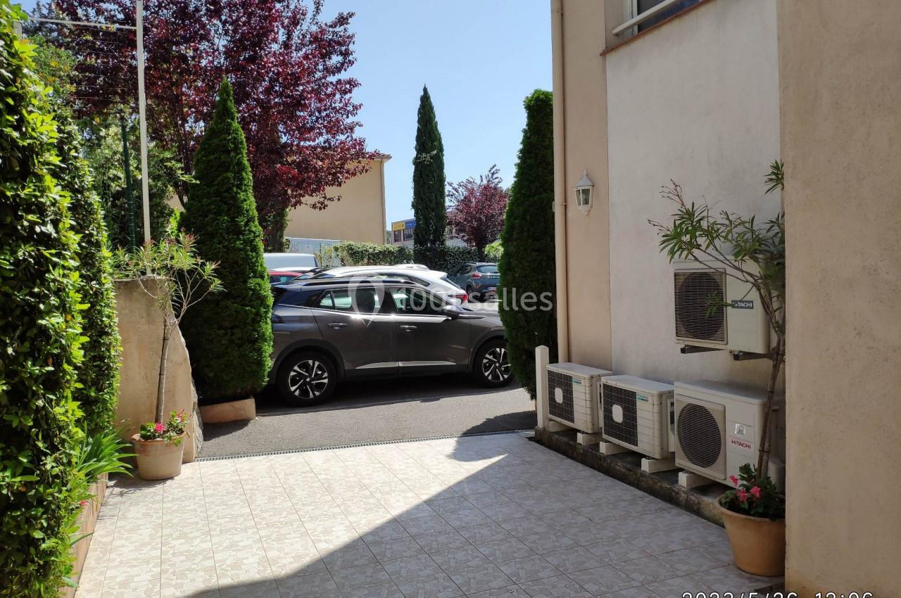 Entrée dallée avec plantes en pot, vue sur un parking avec voitures et arbres en arrière-plan.