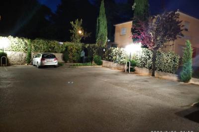 Parking extérieur de nuit avec une voiture stationnée, éclairé par des lampadaires et entouré de haies et d'arbres.