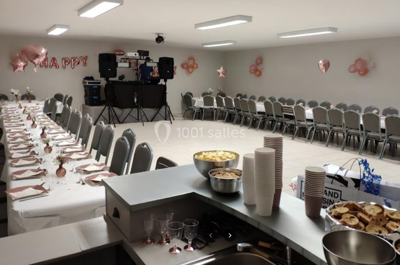 Salle décorée pour une fête avec tables dressées, chaises alignées, ballons roses et espace pour un DJ.