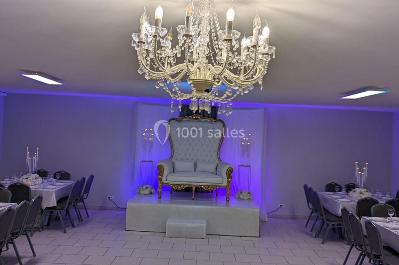 Salle de réception décorée avec un grand lustre, un fauteuil élégant sur une estrade et des tables dressées.
