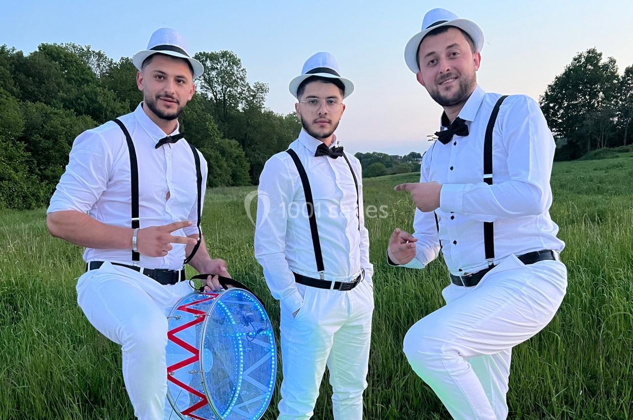Trois hommes en tenue blanche avec chapeaux et bretelles, posant dans un champ verdoyant, l'un tenant un tambour lumineux.