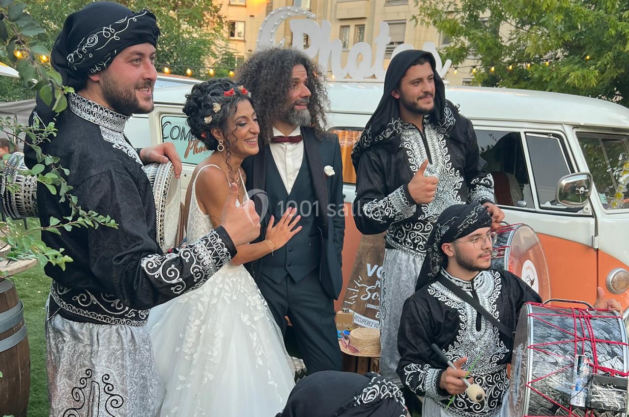 Un couple de mariés pose avec des danseurs en tenue traditionnelle devant un van décoré en extérieur.