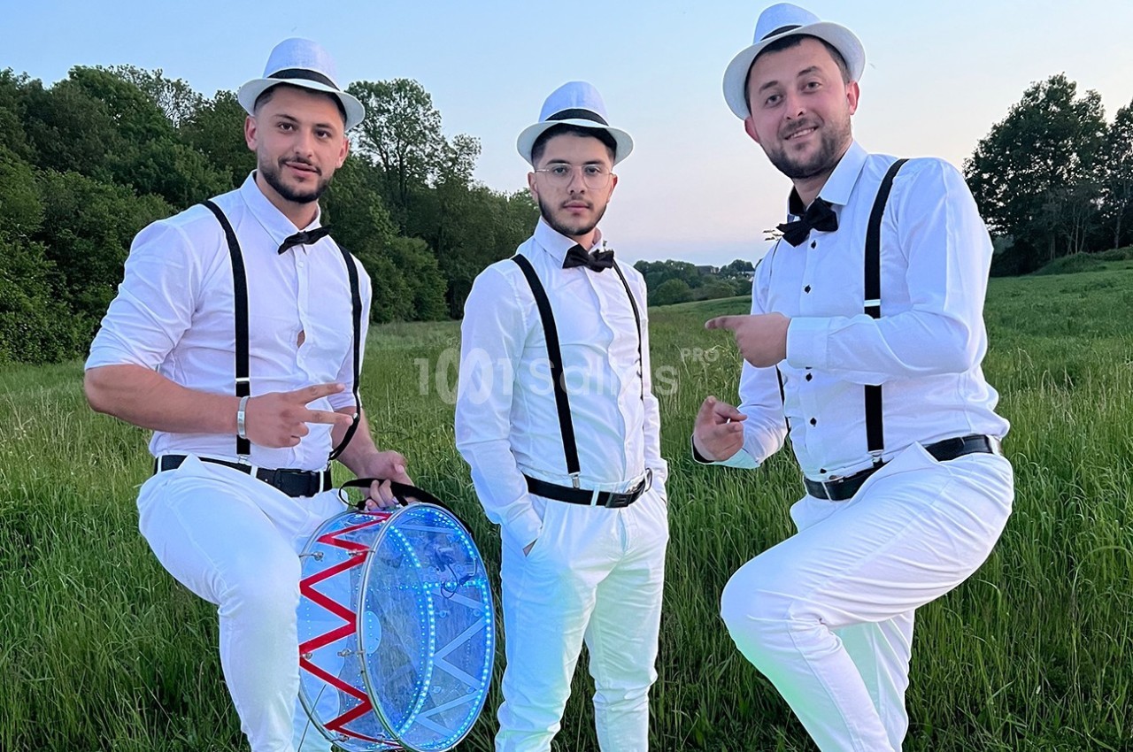 Trois hommes en tenue blanche avec chapeaux et nœuds papillon posent dans un champ, l'un tenant un tambour lumineux.