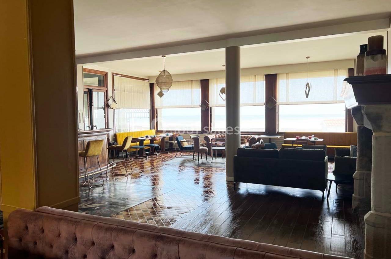 Salle lumineuse avec grandes baies vitrées, mobilier varié en bois et tissu, vue sur l'extérieur en arrière-plan.