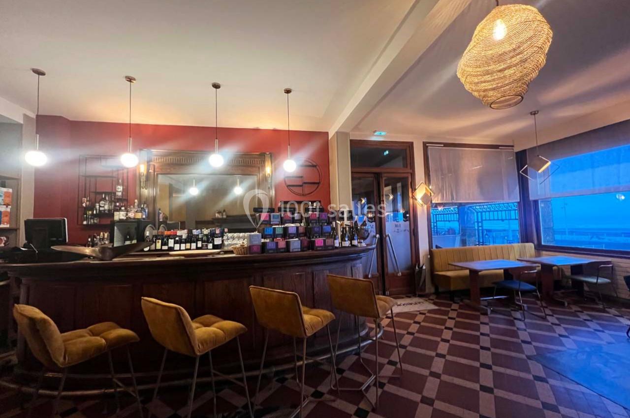 Bar avec comptoir en bois, tabourets jaunes, tables et banquettes, éclairage suspendu et grande fenêtre donnant sur l…