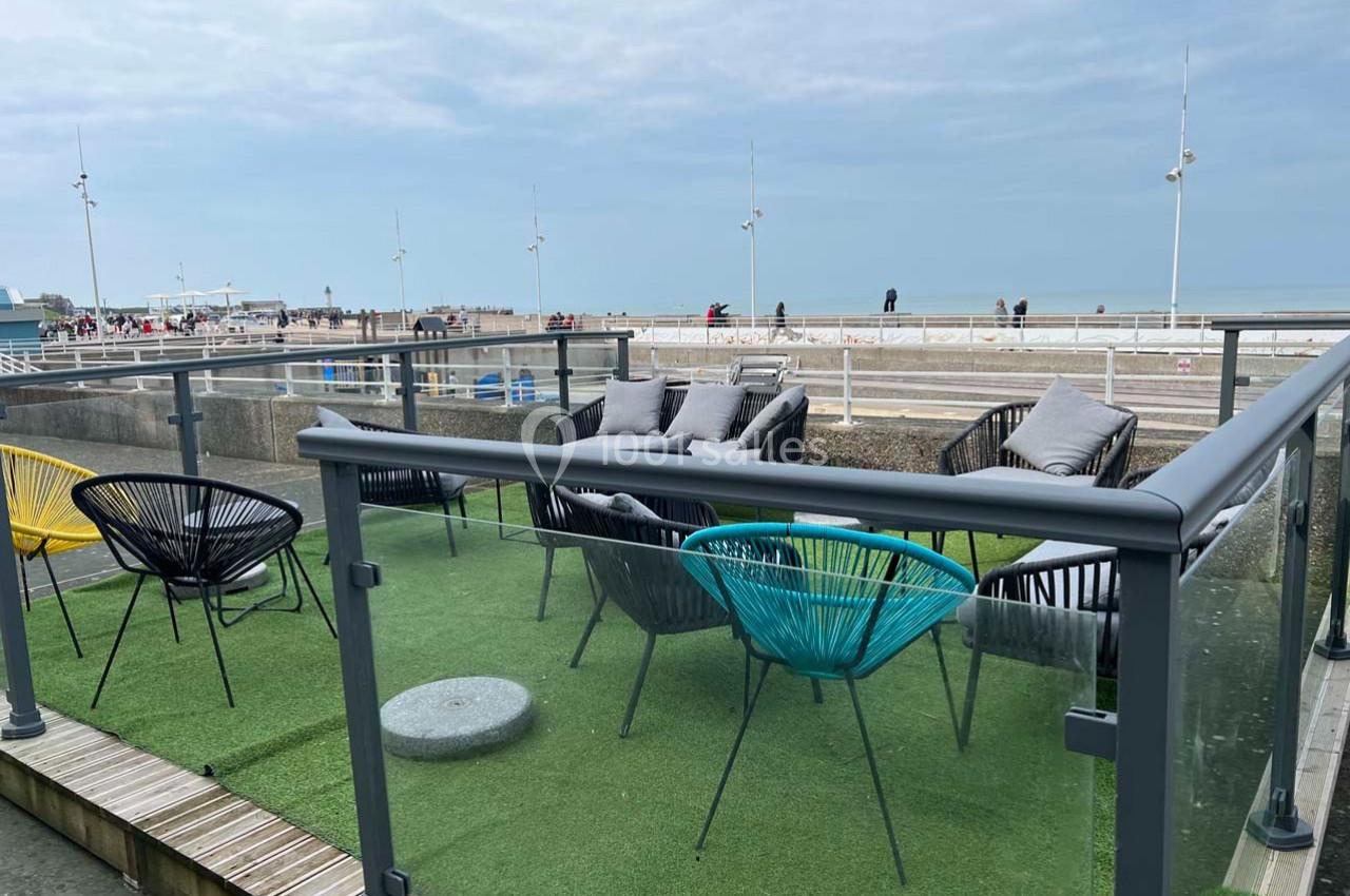 Terrasse avec fauteuils colorés et table basse, vue sur une plage et la mer sous un ciel nuageux.