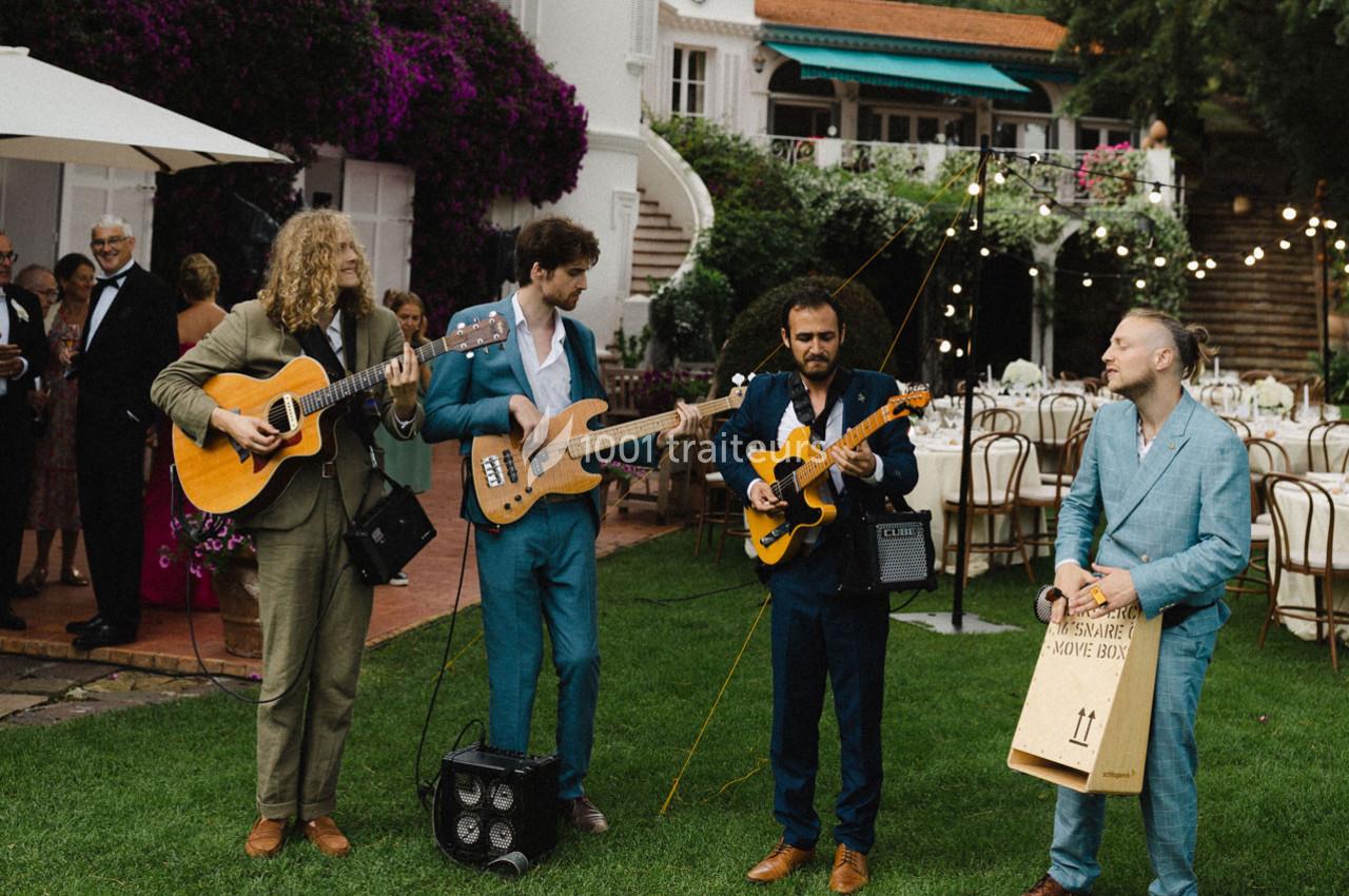 Quatre musiciens jouent en plein air dans un jardin lors d'un événement, entourés de tables et de guirlandes lumineuses.