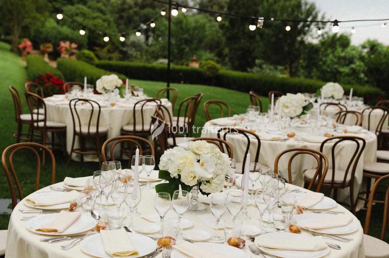 Tables rondes élégamment dressées avec nappes blanches, chaises en bois et guirlandes lumineuses dans un jardin verdoyant.
