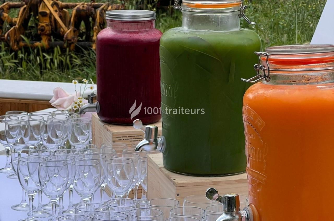 Trois grandes jarres de jus colorés (rouge, vert, orange) avec robinets, entourées de verres sur une table en extérieur.