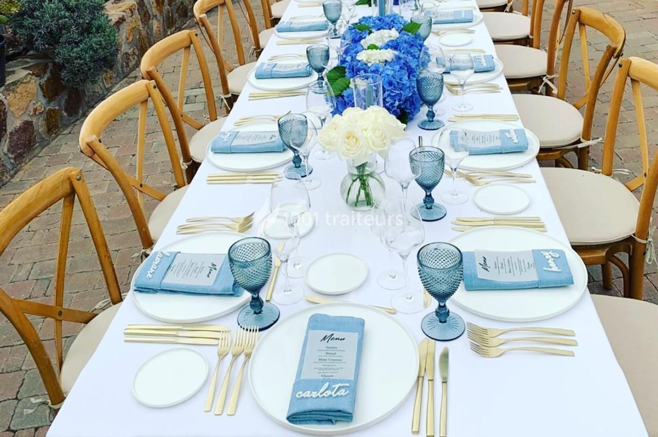 Table dressée pour un repas, avec nappes blanches, serviettes bleues, verres colorés et centre de table floral.