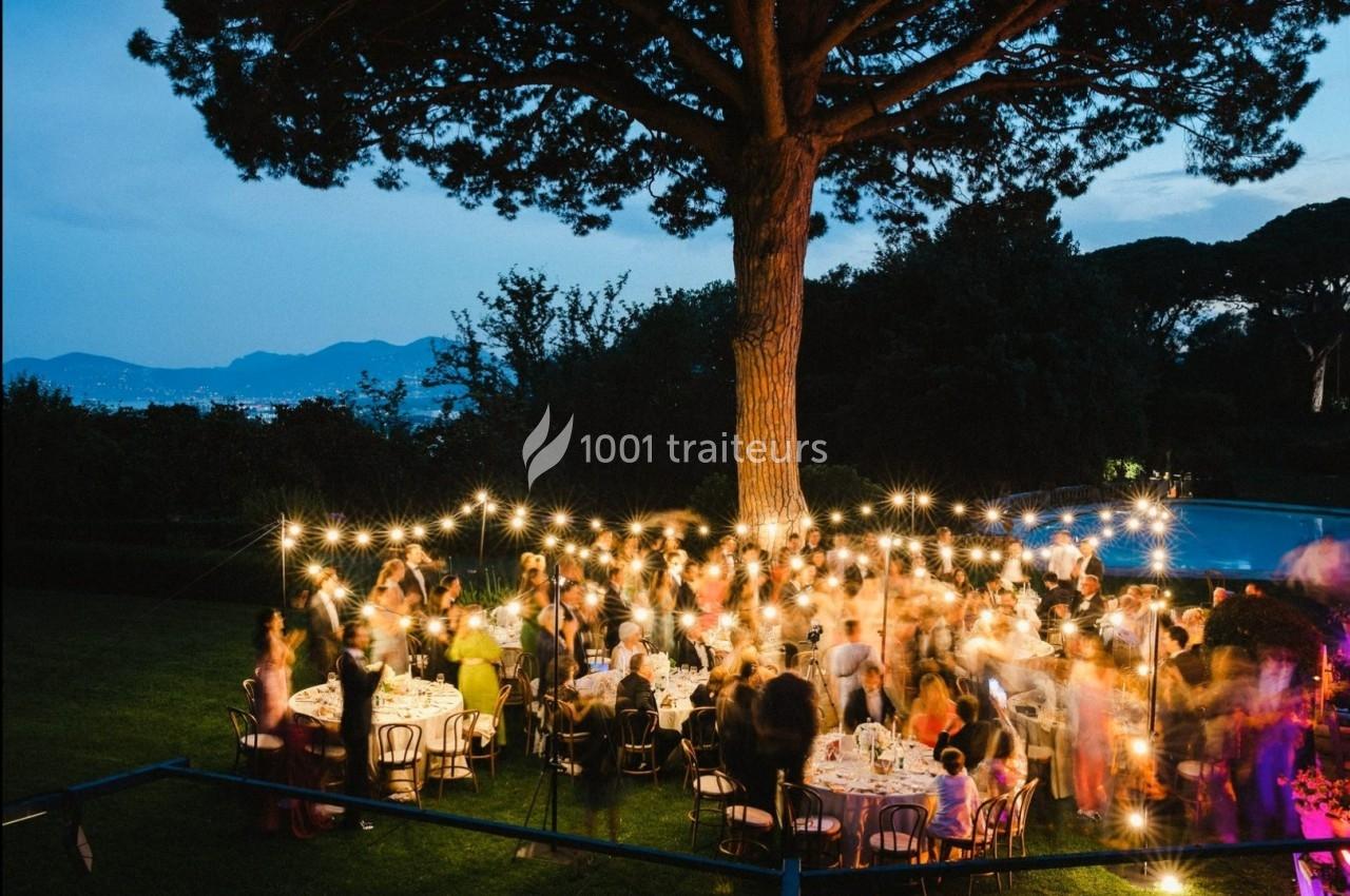 Groupe de personnes dînant en plein air sous des guirlandes lumineuses, entourées d'arbres et d'un paysage au crépuscule.