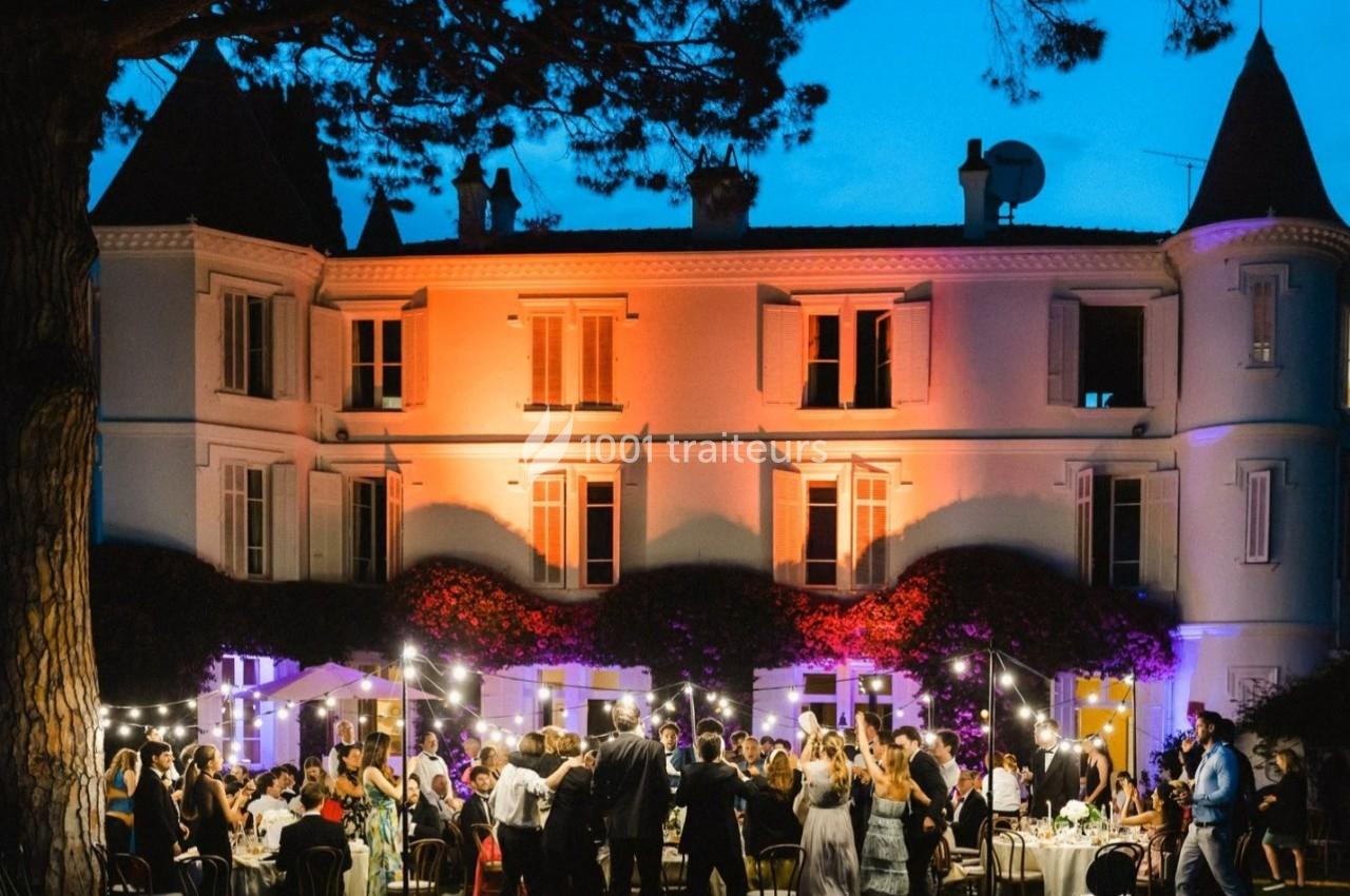 Groupe de personnes dînant en soirée devant une grande maison illuminée, entourée de guirlandes lumineuses.