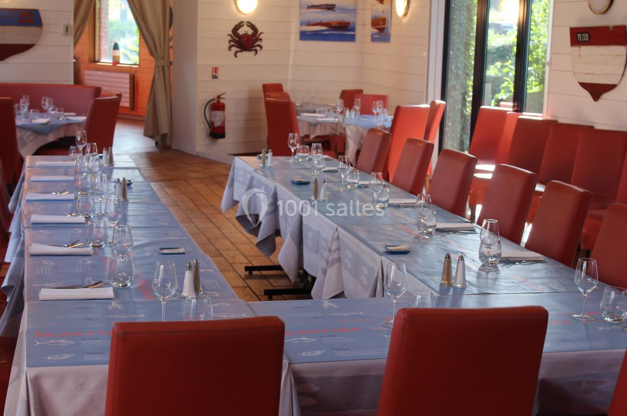 Salle de restaurant avec de longues tables dressées, nappes bleues, chaises rouges et lumière naturelle.