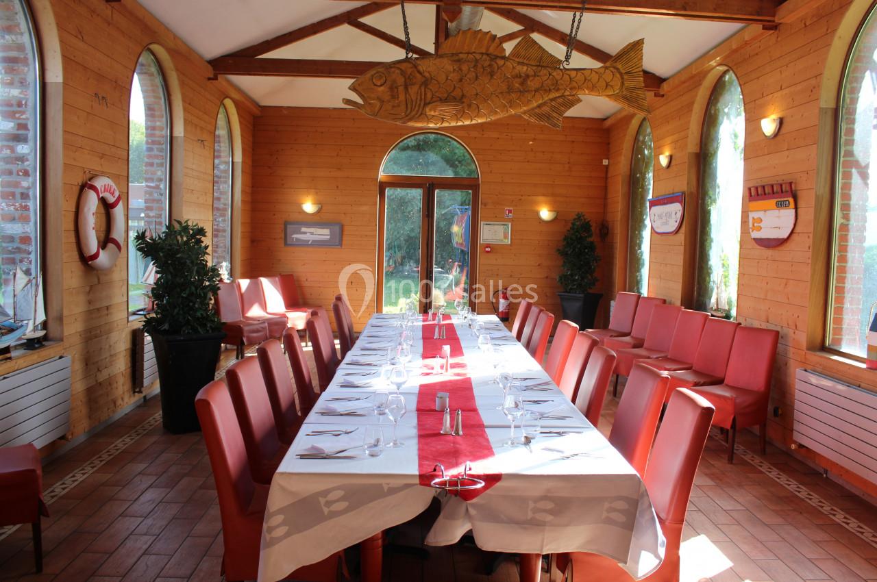 Salle lumineuse avec grande table dressée, chaises rouges, murs en bois, fenêtres cintrées et décoration en forme de poisson.