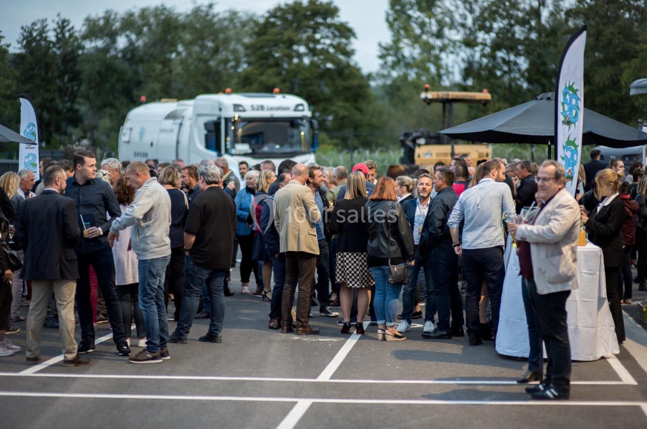 Groupe de personnes rassemblées en extérieur lors d'un événement, avec des camions et des stands en arrière-plan.