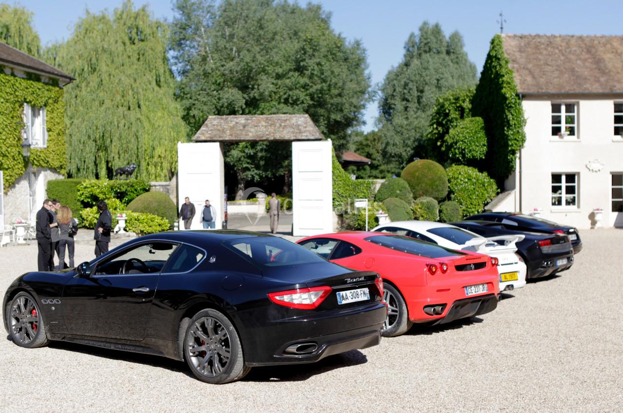 Alignement de voitures de sport luxueuses sur une allée gravillonnée devant un bâtiment entouré de verdure.