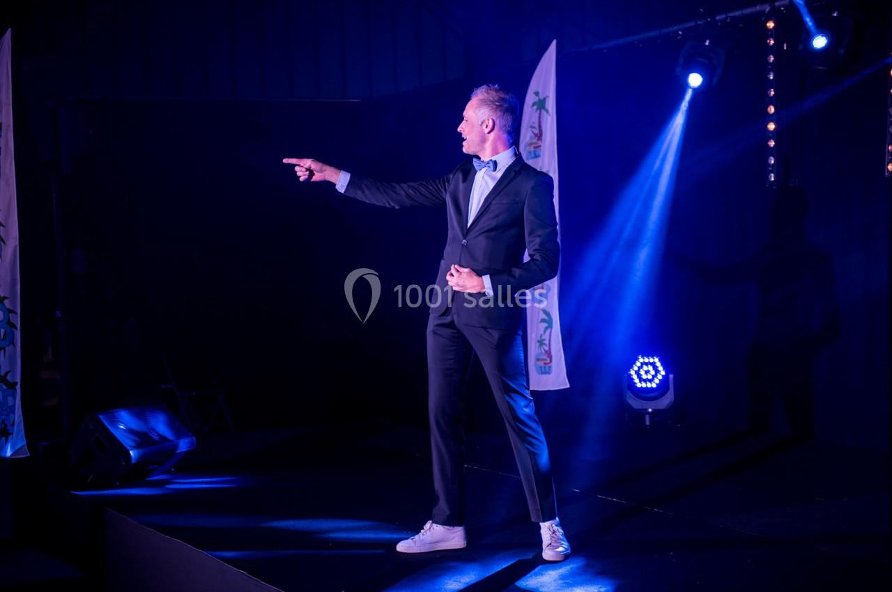 Un homme en costume noir et baskets blanches pointe du doigt sur une scène éclairée par des projecteurs.
