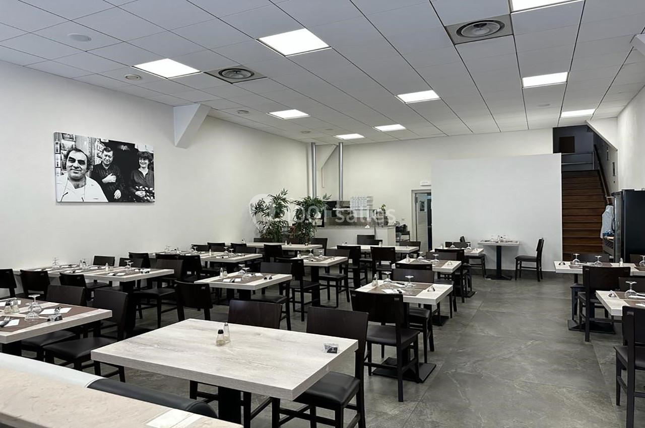Salle de restaurant moderne avec tables dressées, chaises sombres, murs blancs et photo en noir et blanc au mur.