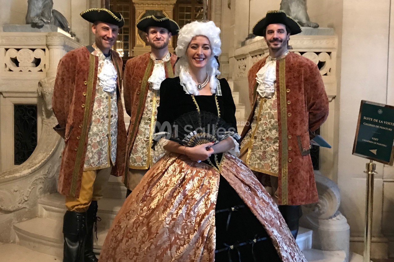 Personnes en costumes d'époque baroque posant dans un décor intérieur élégant avec des escaliers en arrière-plan.