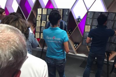 Deux personnes assises dans un buggy bleu en extérieur, discutant et manipulant des objets, avec un homme debout à côté.