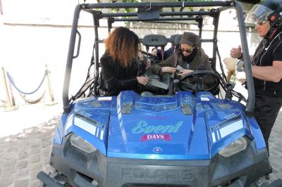Deux personnes assises dans un buggy bleu en extérieur, discutant et manipulant des objets, avec un homme debout à côté.