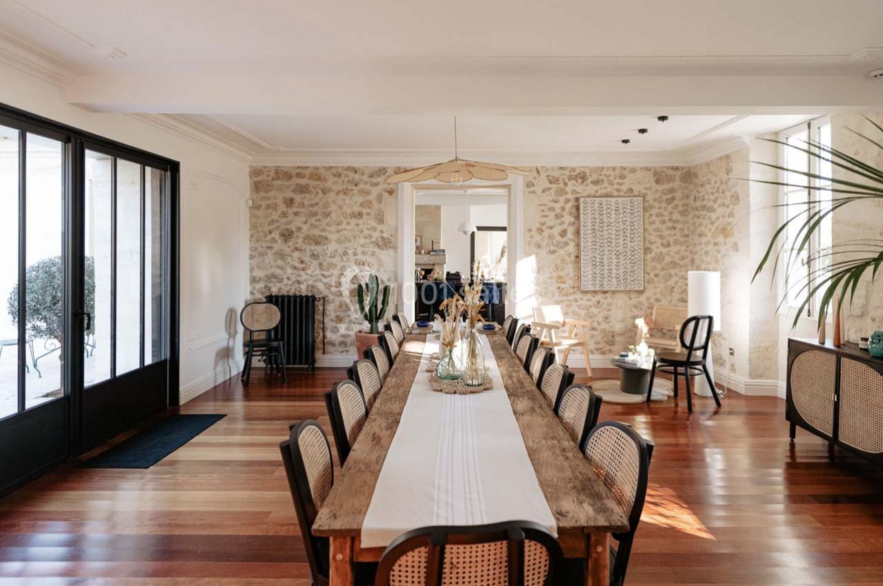 Salle à manger lumineuse avec une longue table en bois, des chaises en rotin, un sol en parquet et des murs en pierre…