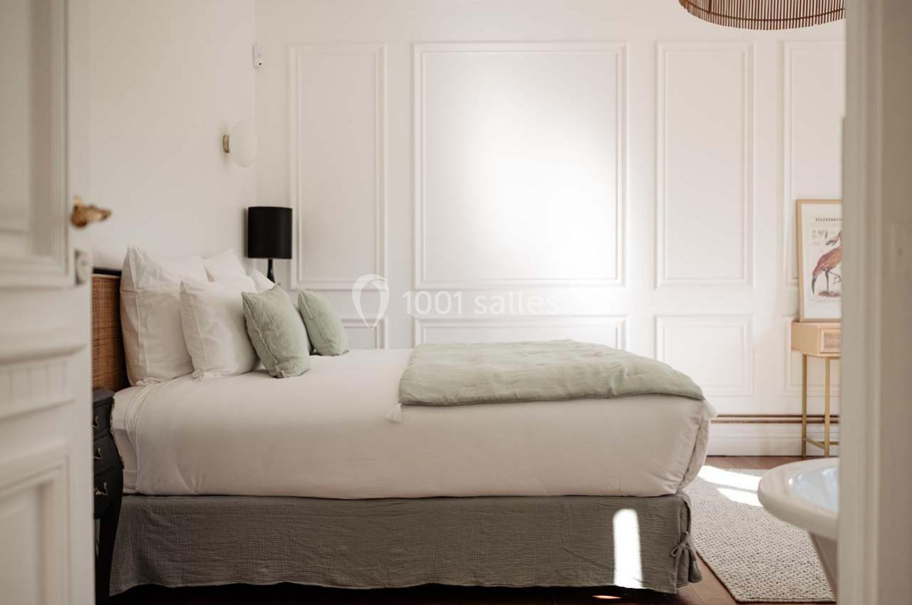 Chambre lumineuse avec un lit double, draps blancs, coussins verts, murs blancs moulurés et mobilier minimaliste.