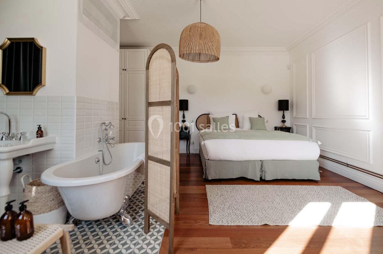 Chambre lumineuse avec lit double, baignoire sur pieds, paravent en rotin et décoration élégante dans des tons neutres.