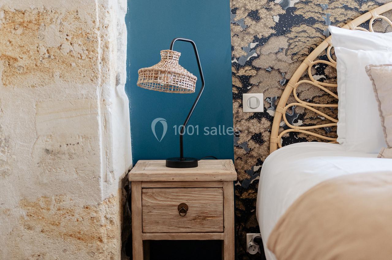 Lampe en rotin sur une table de chevet en bois, près d'un lit avec tête en rotin et mur bleu décoré.