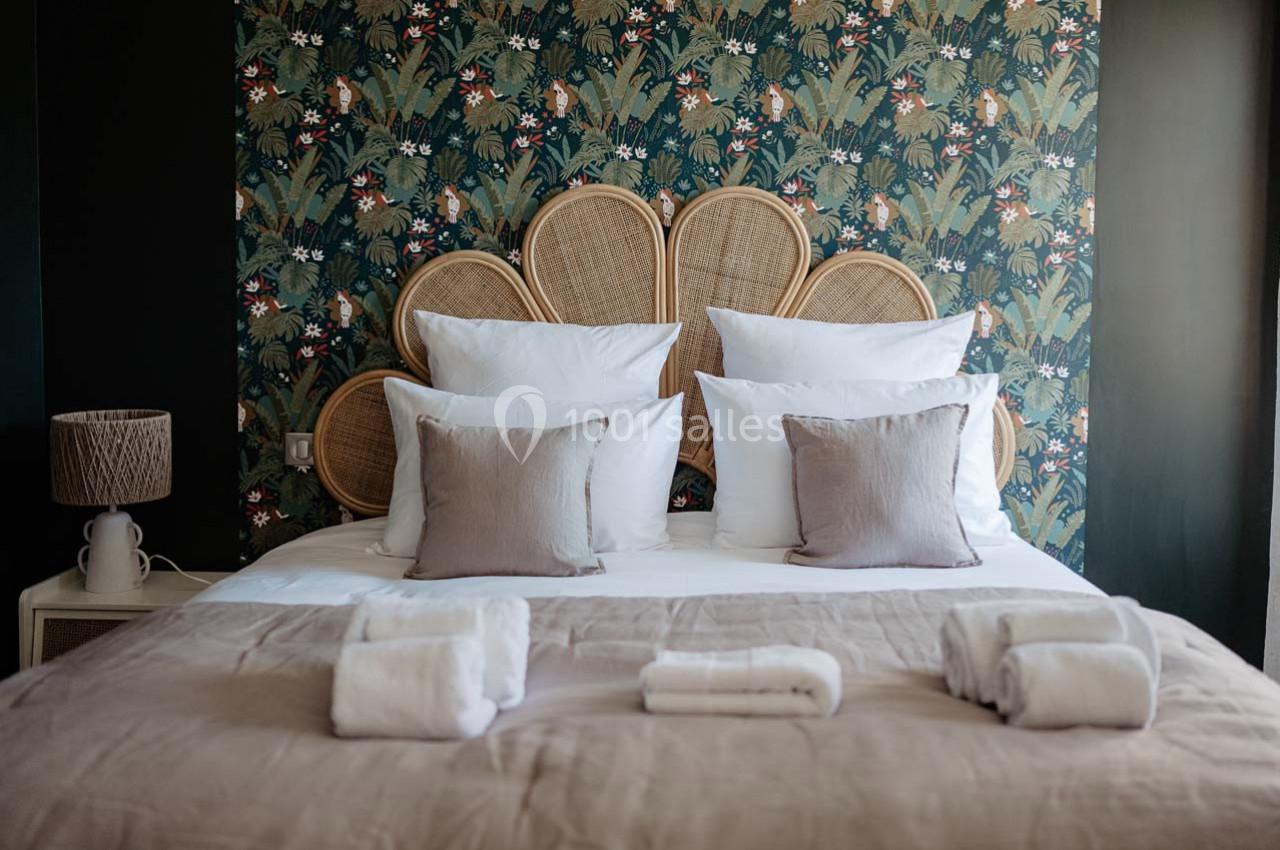 Lit double avec tête de lit en rotin, draps blancs, coussins beiges et fond de papier peint à motifs végétaux.