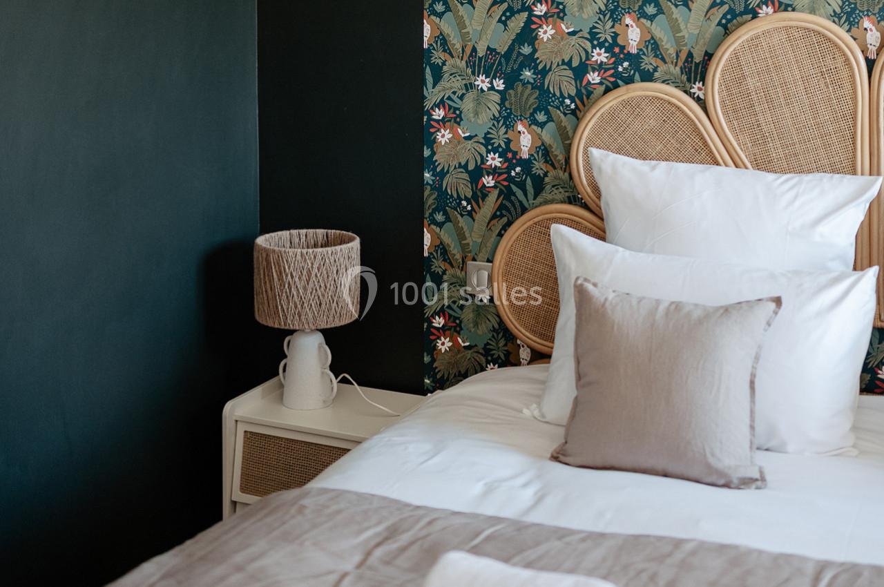 Lit avec coussins blancs et beiges, lampe en rotin sur une table de chevet, mur vert et papier peint floral en arrière-plan.